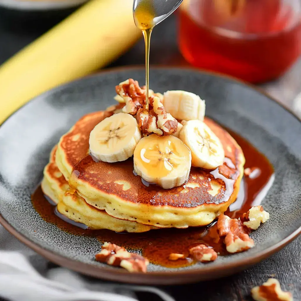 Une assiette avec trois pancakes moelleux garnis de tranches de banane, noix hachées et un filet de sirop.
