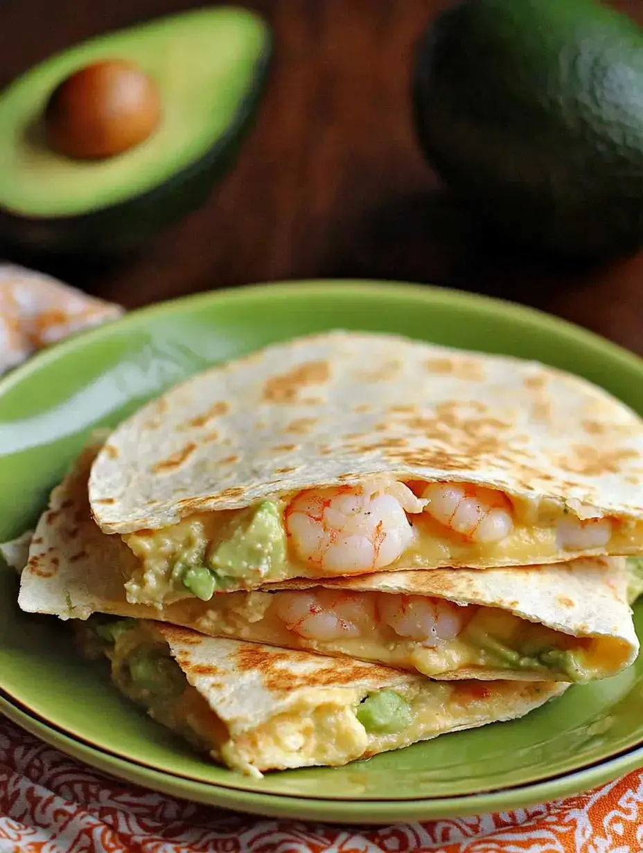Un gros plan d'une assiette verte présentant des quesadillas empilées garnies de crevettes et d'avocat, accompagnées d'un avocat coupé en deux en arrière-plan.