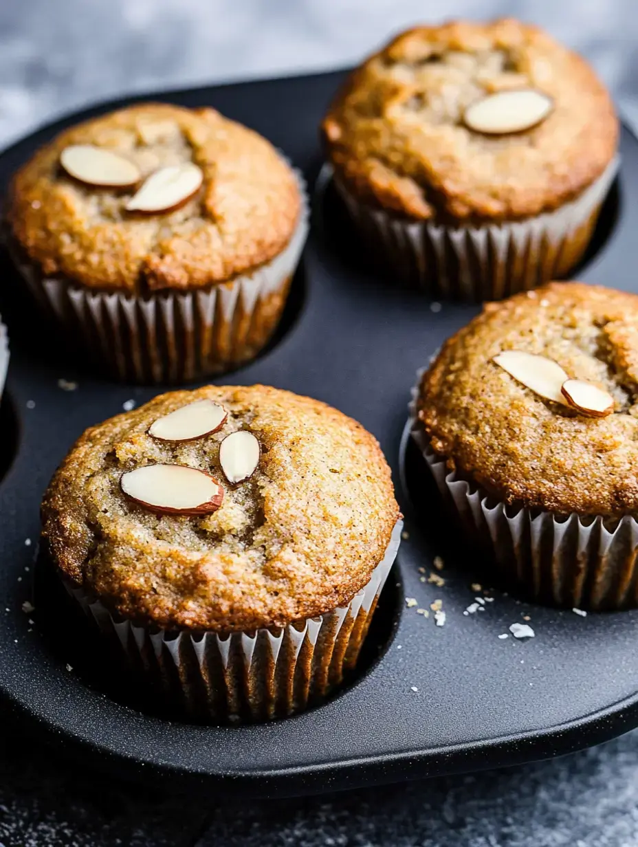 Quatre muffins aux amandes fraîchement cuits placés dans un moule à muffins noir avec des amandes effilées par-dessus.