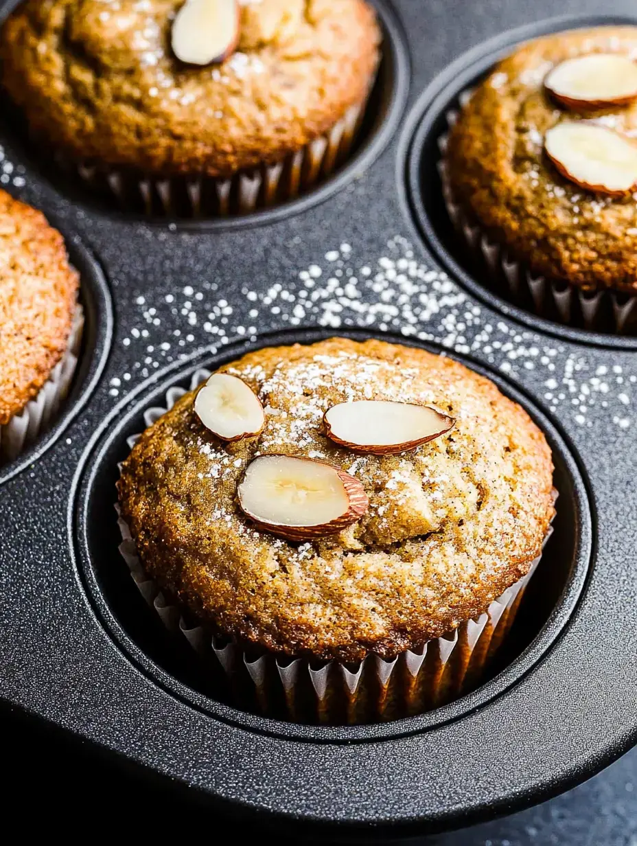 Un gros plan de muffins fraîchement cuits garnis d'amandes effilées et de sucre glace, disposés dans un moule à muffins foncé.