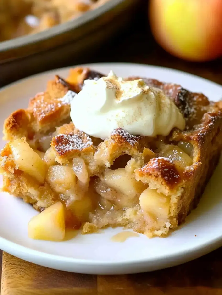Une part de tarte aux pommes servie sur une assiette, surmontée de crème fouettée et saupoudrée de sucre glace, avec une pomme entière en arrière-plan.